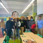 Group photo of Stem Academy Local Leaders with Mr. and Mrs. Ritz of the Green Bronx Machine - blue/green in the background along with a cartoon mural of Mr. Ritz, and bright white tower gardens to the side. In front of them are a few beige desks with bright green chairs.