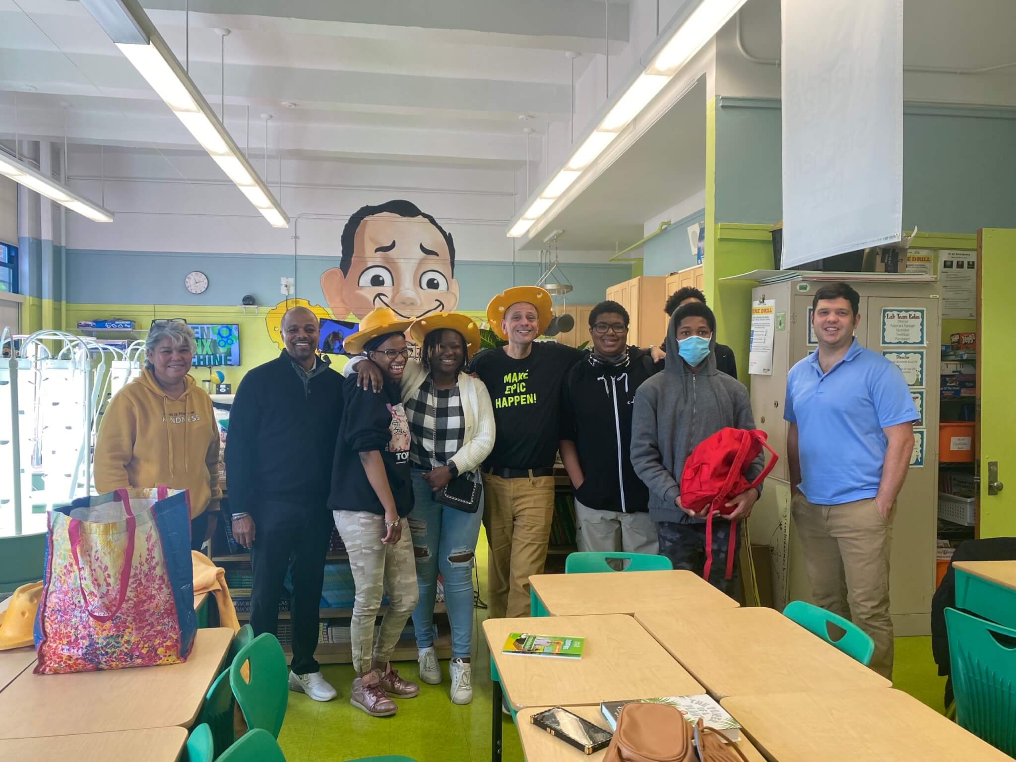 Group photo of Stem Academy Local Leaders with Mr. and Mrs. Ritz of the Green Bronx Machine - blue/green in the background along with a cartoon mural of Mr. Ritz, and bright white tower gardens to the side. In front of them are a few beige desks with bright green chairs.