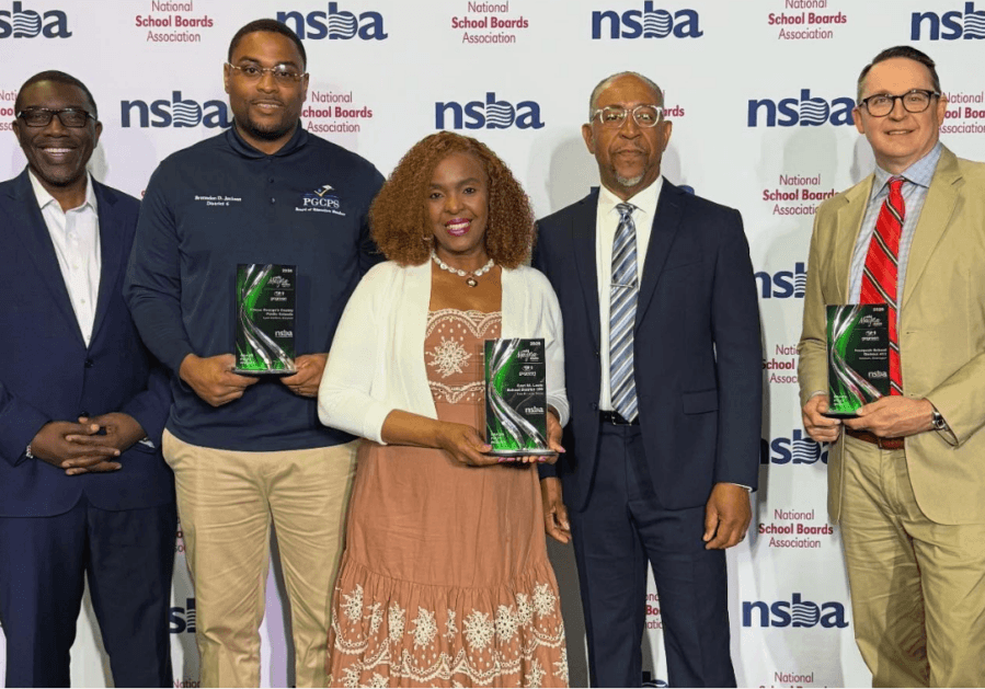 Five people smile on stage holding Magna Awards trophies in front of an NSBA backdrop, celebrating their districts' recognition for healthy, sustainable schools.