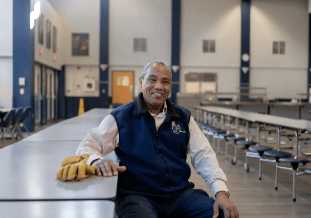 A smiling school facilities manager sits at a cafeteria table, work gloves resting beside him, in an empty school cafeteria.