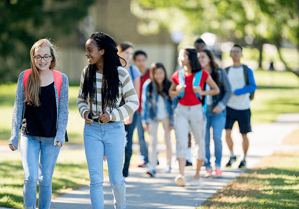 Two teenage girls walk and laugh together in the foreground, backpacks on, while a larger group of middle and high school students follows behind them on a tree-lined path outside a school.