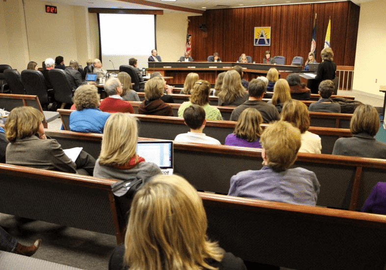 A packed school board meeting room, with board members seated at a raised dais in front of district and state flags, and rows of community members filling the audience seats.
